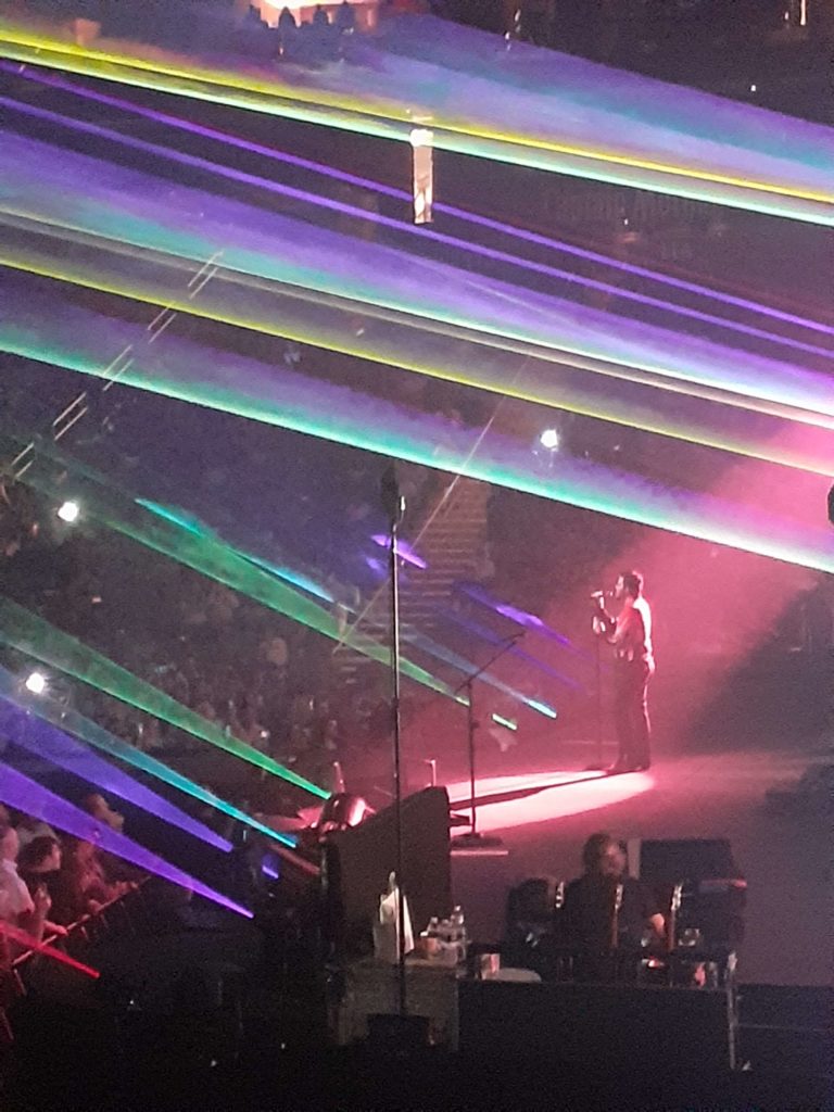 Adam Lambert singing under rainbow spotlights.